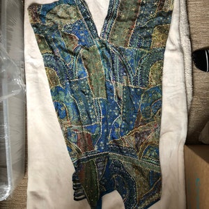 May include: A patchwork jumpsuit in shades of blue, green, and gold, with detailed patterns and embellishments. The halter-style top flows into wide-leg trousers. The fabric looks lightweight, ideal for warmer weather.