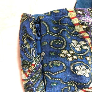 May include: Close-up of a blue fabric with a detailed floral pattern. A blue zip is visible on the side. The fabric has a mix of colours, including green, purple, and red, creating a rich, textured appearance. The image focuses on the details of the textile.