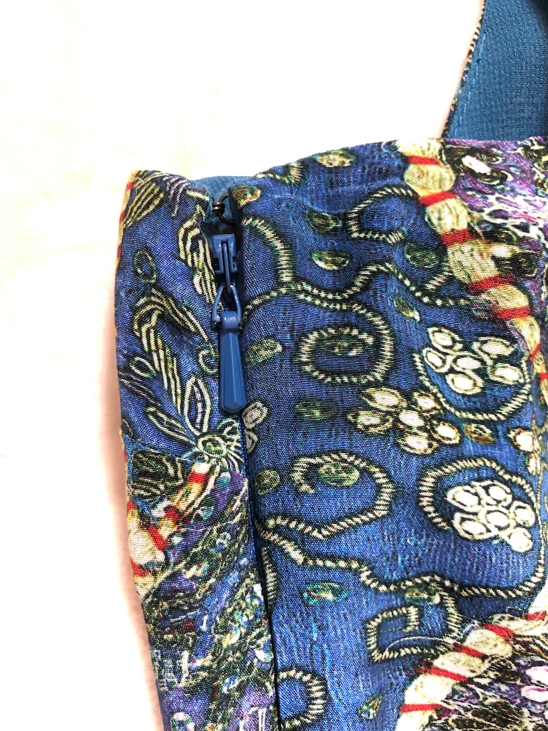 May include: Close-up of a blue fabric with a detailed floral pattern. A blue zip is visible on the side. The fabric has a mix of colours, including green, purple, and red, creating a rich, textured appearance. The image focuses on the details of the textile.