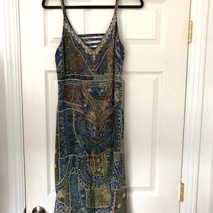 May include: A patterned midi dress with a V-neck and spaghetti straps hangs on a black hanger. The dress features a patchwork design in shades of blue, green, and brown, with intricate detailing and a criss-cross back.