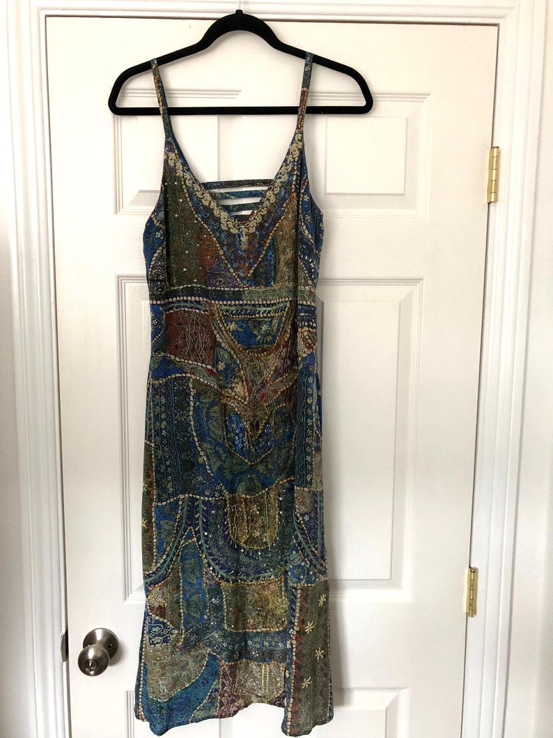 May include: A patterned midi dress with a V-neck and spaghetti straps hangs on a black hanger. The dress features a patchwork design in shades of blue, green, and brown, with intricate detailing and a criss-cross back.