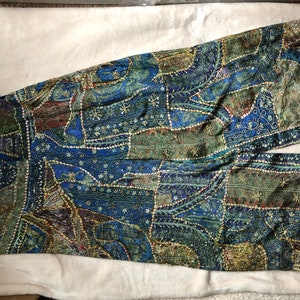 May include: A patchwork garment in shades of blue, green, and brown. The fabric features intricate patterns and is embellished with small, sparkling accents. The garment appears to be wide-legged trousers or a jumpsuit.