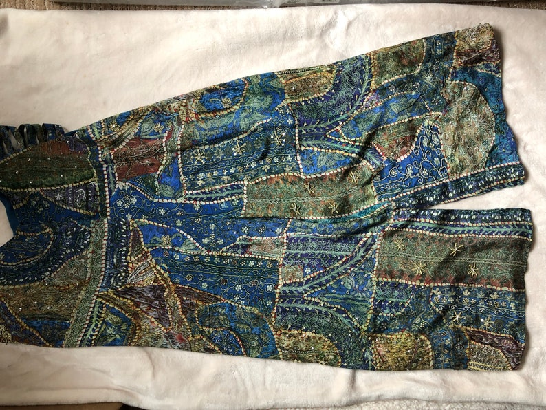 May include: A patchwork garment in shades of blue, green, and brown. The fabric features intricate patterns and is embellished with small, sparkling accents. The garment appears to be wide-legged trousers or a jumpsuit.