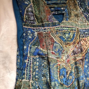 May include: A blue patchwork dress with a V-neck and detailed embroidery. The dress showcases a blend of patterns in blue, green, and brown, with a crisscross design at the neckline. The dress has a side zipper.