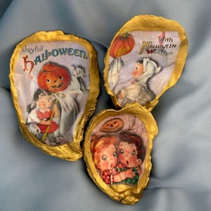 May include: Three vintage-style Halloween oyster shell decorations. Each shell has a gold-painted edge and features a different retro Halloween image with text. The images include a pumpkin, children, and the phrases "Joyful Halloween" and "Halloween Wishes."