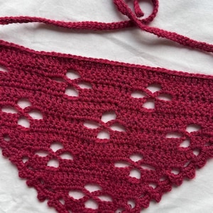 May include: A crocheted burgundy triangle scarf with a delicate openwork pattern. The scarf has a long tie that can be tied around the neck.