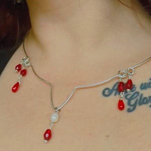 Peut inclure: Un collier ras de cou argenté avec un design en forme de V. Le collier comporte plusieurs perles rouges en forme de larme pendantes et une seule perle blanche.