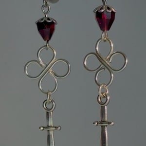 Pode incluir: Um par de brincos de prata com uma conta de cristal vermelho, um nó infinito de prata e um pingente de adaga de prata.