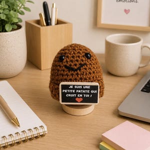 Peut inclure: Une petite patate crochetée de couleur marron avec des yeux en perles noires et un sourire brodé, fixée sur un socle en bois. Elle tient un panneau rectangulaire portant le texte 'JE SUIS UNE PETITE PATATE QUI CROIT EN TOI !' et un cœur rouge.