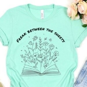 May include: A light green t-shirt with a black and white graphic of a book with flowers growing out of it. The text "Freak Between The Sheets" is printed on the shirt.