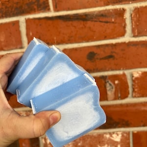 Puede incluir: Una pila de jabones azul claro con bordes ondulados y detalles blancos. El jabón tiene un aspecto texturizado y se sostiene contra una pared de ladrillos rojos. El jabón parece hecho a mano.