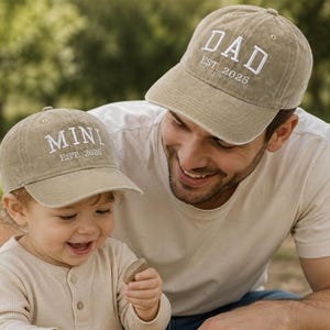 Może przedstawiać: Dwie beżowe czapki z daszkiem, jedna z napisem "DAD" i druga z napisem "MINI", obie z haftem "EST 2025". Czapki noszą uśmiechnięty dorosły i małe dziecko. Tło to rozmyty widok na zewnątrz.