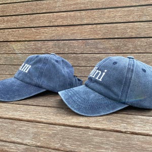 May include: Two blue denim baseball caps with curved visors. One cap has the word "Mum" in white block letters, and the other has "Mini". The caps are on a wooden surface.