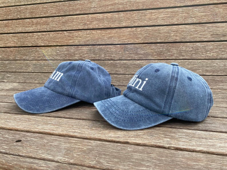 May include: Two blue denim baseball caps with curved visors. One cap has the word "Mum" in white block letters, and the other has "Mini". The caps are on a wooden surface.