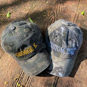 Puede incluir: Dos gorras de b&eacute;isbol de camuflaje con texto bordado. Una gorra tiene el texto "GEORGE K" en hilo amarillo. La otra gorra tiene el texto "CUSTOM TEXT" en hilo blanco.