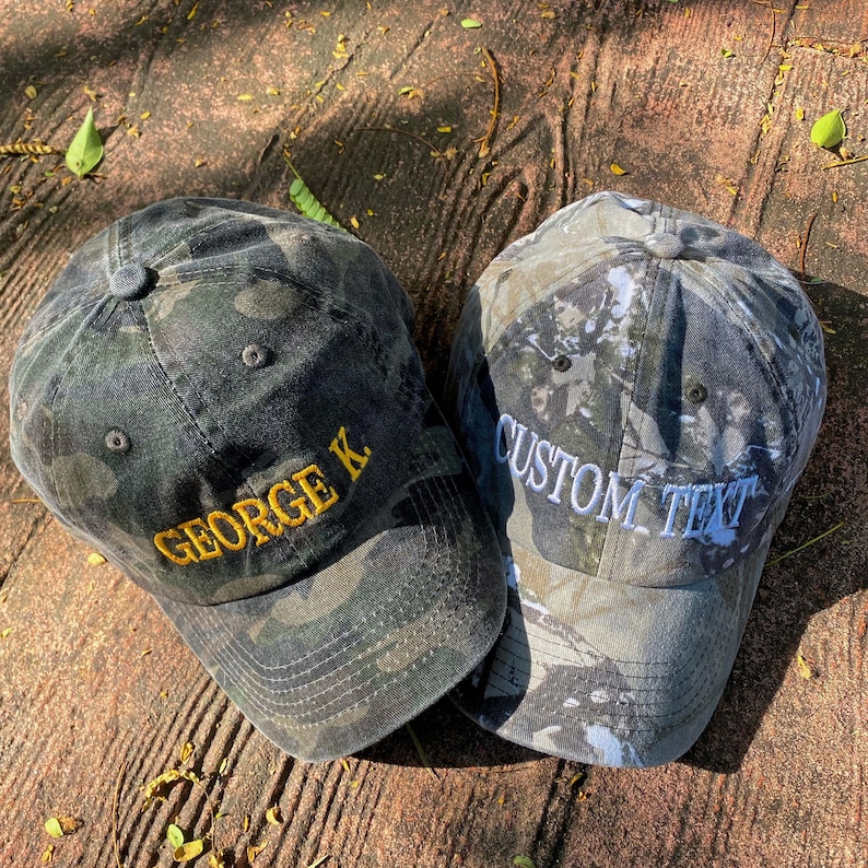Puede incluir: Dos gorras de b&eacute;isbol de camuflaje con texto bordado. Una gorra tiene el texto "GEORGE K" en hilo amarillo. La otra gorra tiene el texto "CUSTOM TEXT" en hilo blanco.
