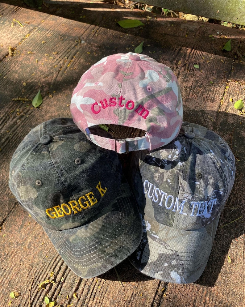 Puede incluir: Tres gorras de b&eacute;isbol de camuflaje con texto bordado. Una gorra es rosa con camuflaje blanco y dice "Custom" en rosa. Una gorra es negra con camuflaje verde y dice "GEORGE K" en amarillo. Una gorra es gris con camuflaje negro y dice "CUSTOM TEXT" en blanco.