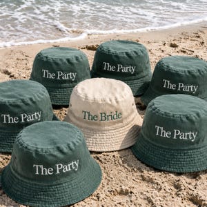 Op de afbeelding: Een verzameling van zeven bucket hats gerangschikt op zand. Zes hoeden zijn donkergroen met de witte geborduurde tekst "The Party" aan de voorzijde. Eén centrale hoed is crèmekleurig met de donkergroene geborduurde tekst "The Bride".