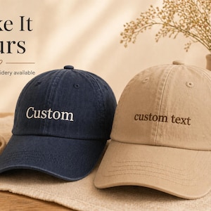 May include: Two baseball caps, one navy blue and one tan, are displayed. The navy cap has "Custom" embroidered in white, while the tan cap has "custom text" embroidered in a matching tan color. The image includes the text "Make It Yours" and "Custom embroidery available."