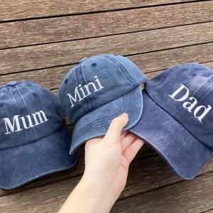 May include: Three blue denim baseball caps with white embroidered text. The caps read "Mum", "Mini", and "Dad". The caps are displayed on a wooden surface.