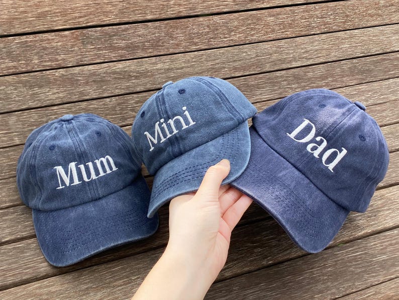 May include: Three blue denim baseball caps with white embroidered text. The caps read "Mum", "Mini", and "Dad". The caps are displayed on a wooden surface.