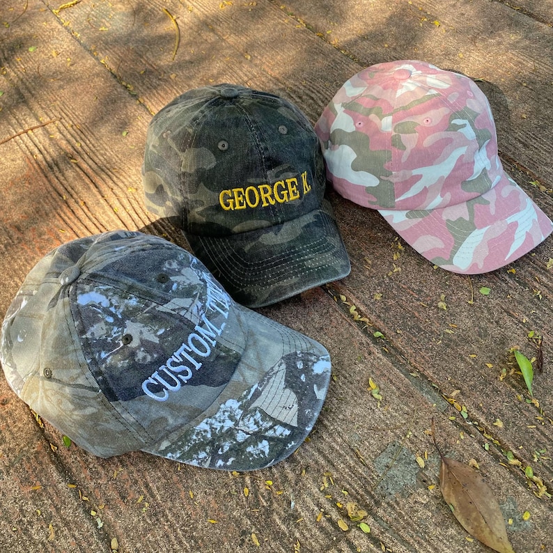 Puede incluir: Tres gorras de b&eacute;isbol de camuflaje. Una es negra y verde con el texto "GEORGE K" bordado en amarillo. Otra es de camuflaje rosa y verde. La tercera es un camuflaje verde oscuro y gris con el texto "CUSTOM TOM" bordado en blanco.