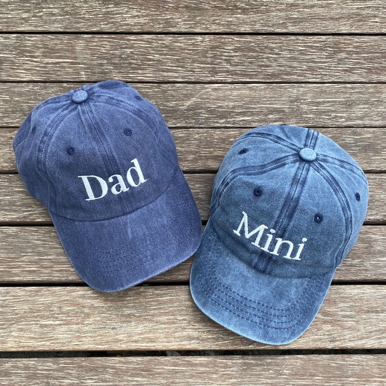 May include: Two blue baseball caps with white text. One cap says "Dad", the other says "Mini". The caps are made of a soft material and have a classic, vintage look. Perfect for matching outfits.