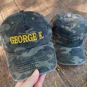 Puede incluir: Dos gorras de b&eacute;isbol de camuflaje, una con el nombre "GEORGE K." bordado en amarillo en la parte delantera.