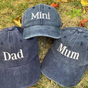 Puede incluir: Tres gorras de béisbol azul marino con texto bordado en blanco. Una gorra dice "Mini", otra dice "Dad" y la tercera dice "Mum". Las gorras están dispuestas sobre un lecho de hierba verde y hojas caídas.