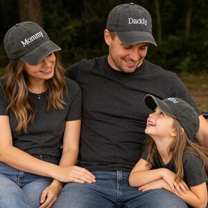 May include: A family of three wearing matching gray baseball caps and t-shirts. The caps are embroidered with the words "Mommy", "Daddy", and "Mini". The family is sitting outside, smiling and looking at each other.