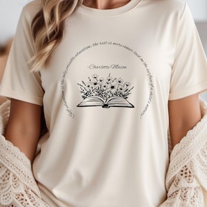 May include: Cream-colored t-shirt featuring a black graphic of an open book with flowers and text. The text reads "Self-education is the only possible education: the rest is more veneer laid on the surface of a child's nature." and "-Charlotte Mason."