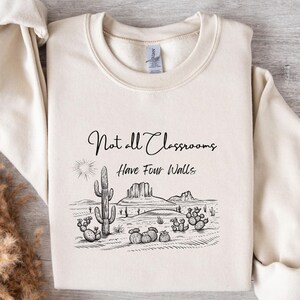Puede incluir: Sudadera de color crema con un gráfico de paisaje desértico. El gráfico presenta un cactus, montañas y el texto "Not all Classrooms Have Four Walls" en escritura negra. La sudadera tiene cuello redondo y mangas largas.