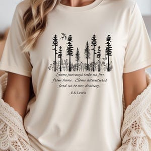 May include: Cream-colored t-shirt featuring a black graphic of stylized trees, flowers, and a bird. The shirt includes a quote by C.S. Lewis: "Some journeys take us far from home. Some adventures lead us to our destiny."