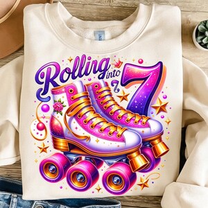 Könnte beinhalten: Ein cremefarbenes Sweatshirt mit einer Grafik von zwei weißen und pinken Rollschuhen mit goldenen Schnürsenkeln und Rollen. Der Text "Rolling into 7" ist in Lila und Pink über den Rollschuhen gedruckt, umgeben von goldenen Sternen, pinken Kugeln und einer Rose.