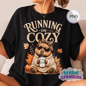 Può includere: Una t-shirt nera con la grafica di un capibara che indossa occhiali da sole e un fiocco leopardato, vestito con un maglione beige. Il capibara tiene in mano una tazza di caffè con la scritta "Fueled by Chaos" e un'icona di zucca. Scritta "RUNNING ON COZY".