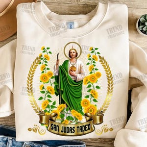 Puede incluir: Sudadera color crema con un diseño de San Judas Tadeo, rodeado de girasoles y una corona de laurel dorada. El diseño incluye el texto "SAN JUDAS TADEO" en una pancarta dorada. La sudadera tiene cuello redondo y mangas largas.
