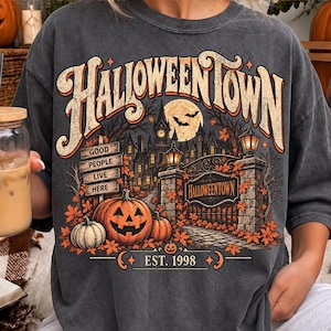 Puede incluir: Una camiseta de manga corta gris carbón con un estampado gráfico de una mansión espeluznante, calabazas y hojas de otoño. El texto dice HALLOWEENTOWN, GOOD PEOPLE LIVE HERE y EST. 1998. El diseño incluye una luna llena con murciélagos.
