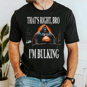 Puede incluir: Una camiseta negra de manga corta con un gráfico de una figura encapuchada con cara de calavera levantando una barra. La figura tiene una gran barriga expuesta. El texto dice "THAT'S RIGHT, BRO" arriba y "I'M BULKING" debajo de la figura.
