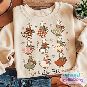 Puede incluir: Una sudadera beige de cuello redondo con un estampado gráfico de nueve patos de dibujos animados con bufandas y tazas de café. Los patos tienen varios patrones, incluyendo leopardo, cuadros y lunares. El texto Hello Fall está impreso debajo.