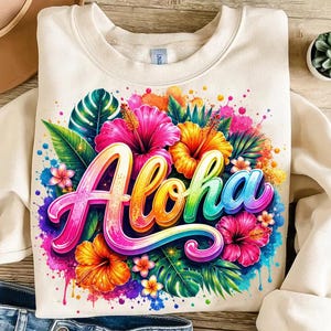May include: A cream-colored crewneck sweatshirt featuring a vibrant, multi-colored graphic print. The design displays the word Aloha in a stylized, rainbow-gradient script, surrounded by colorful hibiscus flowers and tropical green leaves.