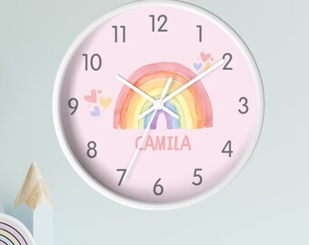 May include: A pink and white round clock with a rainbow and hearts design. The clock has black numbers and a white background. The name "CAMILA" is printed on the clock.