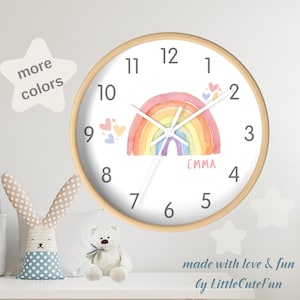May include: A white and wood-framed clock with a rainbow design and the name "EMMA" on the face. The clock has a white background with pink, orange, yellow, green, blue, and purple watercolor rainbow. The clock is surrounded by small pink hearts.