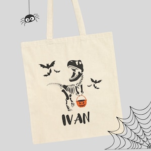 Puede incluir: Bolso tote de color crema con un diseño de esqueleto de dinosaurio negro sosteniendo un cubo de calabaza naranja. Murciélagos negros vuelan alrededor del dinosaurio. El nombre "IVAN" está impreso en negro debajo. Una araña y una telaraña están en el fondo.