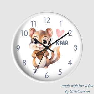 May include: White round wall clock featuring a watercolor illustration of a cute squirrel holding an acorn. The clock face has black hands, gray numerals, and the name "KAIA" with a pink heart. Text at the bottom reads "made with love & fun by LittleCuteFun."