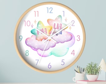 May include: A white clock with a wooden frame features a rainbow watercolor design of clouds and stars. The numbers 1 through 12 are printed in purple on the clock face. The words "Mia" and "Lia" are written on the stars.