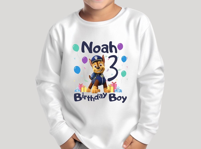 May include: White long-sleeved shirt featuring a cartoon dog in a police uniform. The shirt has the text "Noah 3 Birthday Boy" and colourful balloons.
