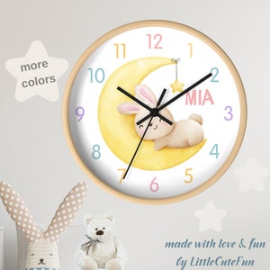May include: A round wall clock with a light wood frame. The clock face features a cartoon bunny on a yellow crescent moon, with the name "MIA" in pink. The clock hands are black, and the numbers are in pastel colors. The text "more colors" is in a star-shaped bubble.
