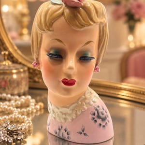 May include: A ceramic head vase depicting a person with blonde hair, a pink rose hair accessory, and a light blue leaf. The figure features closed eyes with black eyelashes, red lips, pink rose earrings, a white lace-style collar, and a pink base.