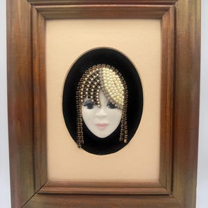 May include: A framed wall art piece featuring a three-dimensional ceramic face mask with a stylized hairstyle made of dark brown and cream-colored beads, clear rhinestones, and faux pearls, set against a black oval background in a wooden frame.