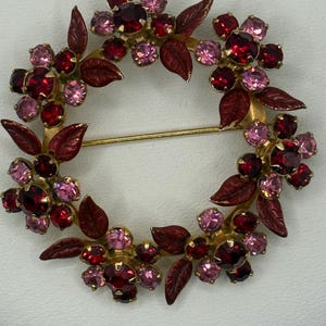 May include: A circular brooch featuring a wreath design composed of dark red and light pink faceted rhinestones set in gold-tone metal. The piece includes small, dark red enameled leaves interspersed among the floral clusters and a pin back.